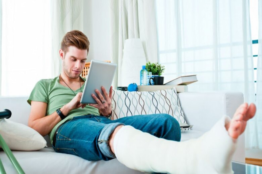 Man with leg cast on couch, using tablet. Discover how to claim and prevent serious injuries in daily life